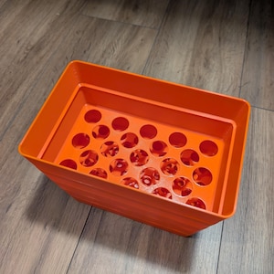 Peut inclure: Un bac de rangement rectangulaire en plastique orange vif doté d'une grille de trous circulaires sur sa base. Le récipient présente des parois lisses et solides ainsi qu'une large ouverture supérieure, avec une structure étagée légèrement effilée.