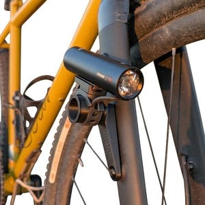 Op de afbeelding: Een zwarte cilindrische fietslamp gemonteerd op de voorvork van een mosterdgele fiets. De lamp heeft een ronde lens en een zwarte kunststof montagebeugel die het apparaat aan het frame bevestigt bij de voorband.
