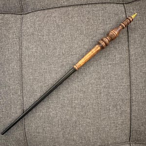 May include: A handcrafted wooden wand with a dark brown handle and a black shaft. The handle features intricate carved details and a gold-colored tip. The wand is approximately 14 inches long.