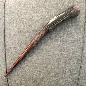 May include: A wooden wand with a dark brown finish, featuring intricate carved details. The wand tapers to a point, with a textured handle and a smooth, polished surface. The wand is approximately 13 inches long.