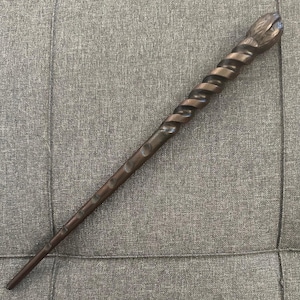 May include: A dark brown wand with a spiraling handle and a detailed tip. The wand has a series of small, evenly spaced indentations along its length. The handle has a twisted design, and the tip has a carved, textured appearance.