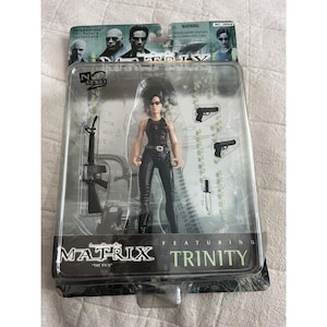 May include: A Trinity action figure from the Matrix film, packaged in a clear plastic case. The figure is wearing a black outfit and sunglasses, with accessories including guns and a knife. The packaging features the film's logo.