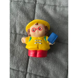 May include: A yellow plastic toy figure of a child wearing a yellow hat, glasses, and a yellow jacket with red pants. The figure is holding a blue book. The toy is designed for children.