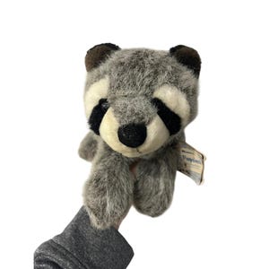 May include: A plush raccoon toy with gray and white fur, black eye markings, and a black nose. The stuffed animal has a tag attached. The toy is held up against a white background.