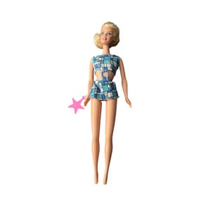 May include: A blonde doll wearing a blue, white, and teal checkered outfit with a matching skirt and a pink star-shaped accessory. The doll has a classic hairstyle and is posed against a white background.