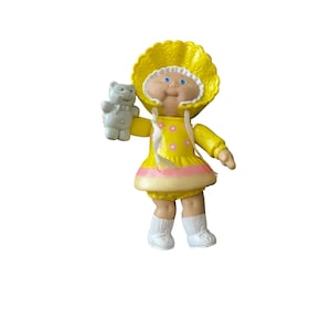 May include: A vintage Cabbage Patch Kids doll wearing a yellow dress with pink accents and a matching bonnet. The doll is holding a small gray teddy bear. The doll has blue eyes and a smiling expression. The doll is wearing white socks and shoes.