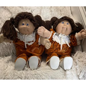 May include: Two Cabbage Patch Kids dolls with brown yarn hair styled in pigtails, blue eyes, and soft fabric bodies. Each doll wears a rust-colored velvet dress with a white lace-trimmed collar, a white ribbon tie, and white shoes.