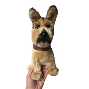 May include: A plush dog toy with tan and black fur, brown leather collar with text. The toy has large, upright ears and is sitting upright. The dog's face has black markings around the eyes and muzzle.