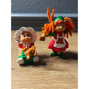 May include: Two vintage Cabbage Patch Kids figurines. One doll wears a red dress and white bonnet, holding a brown teddy bear on a green and white seat. The other doll has orange yarn hair, a green hat, and a red dress with a white apron.