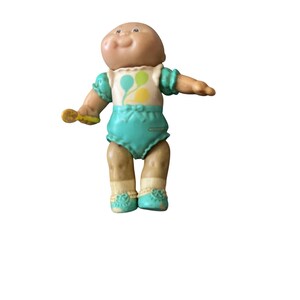May include: A vintage Cabbage Patch Kids doll with a bald head, wearing a teal outfit with a white top featuring balloon graphics. The doll has tan skin, white socks, and teal shoes. It holds a yellow rattle.