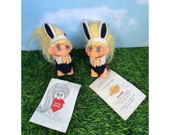 May include: Two identical troll dolls with pale orange skin, blue eyes, and long yellow hair. Each wears a white headband with black bunny ears and a black outfit. Two white paper tags are included, one featuring a drawing of a troll holding a red heart with the text Lucky Troll, and the other with printed manufacturing information.