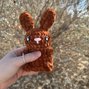 May include: A small brown crocheted bunny with large black eyes and a pink nose. The bunny is being held in a hand.