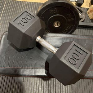 May include: A black, 100-pound dumbbell with a silver handle rests on a black padded bench. The dumbbell has a hexagonal shape with the number "100" embossed on each end. Other weights and a weight rack are in the background.