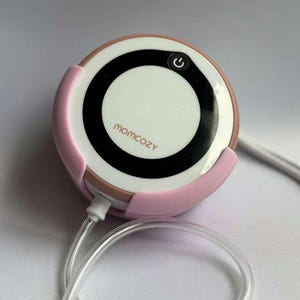 May include: A pink and white breast pump with a black power button and the brand name "momcozy" printed on the front. The pump has a clear tube attached and a light pink cradle.