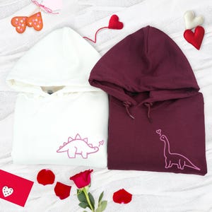 May include: A white hoodie featuring an embroidered pink stegosaurus and a maroon hoodie featuring an embroidered pink long-necked dinosaur, both with matching drawstrings and hoods, displayed alongside decorative fabric hearts and a red rose.