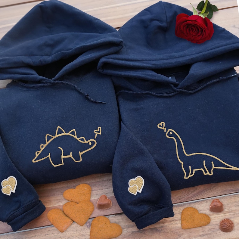 May include: Two navy blue hooded sweatshirts featuring gold embroidered dinosaur outlines on the chest. One hoodie displays a stegosaurus and the other a long-necked dinosaur, each with a small heart. Both sleeves feature two small gold heart patches.