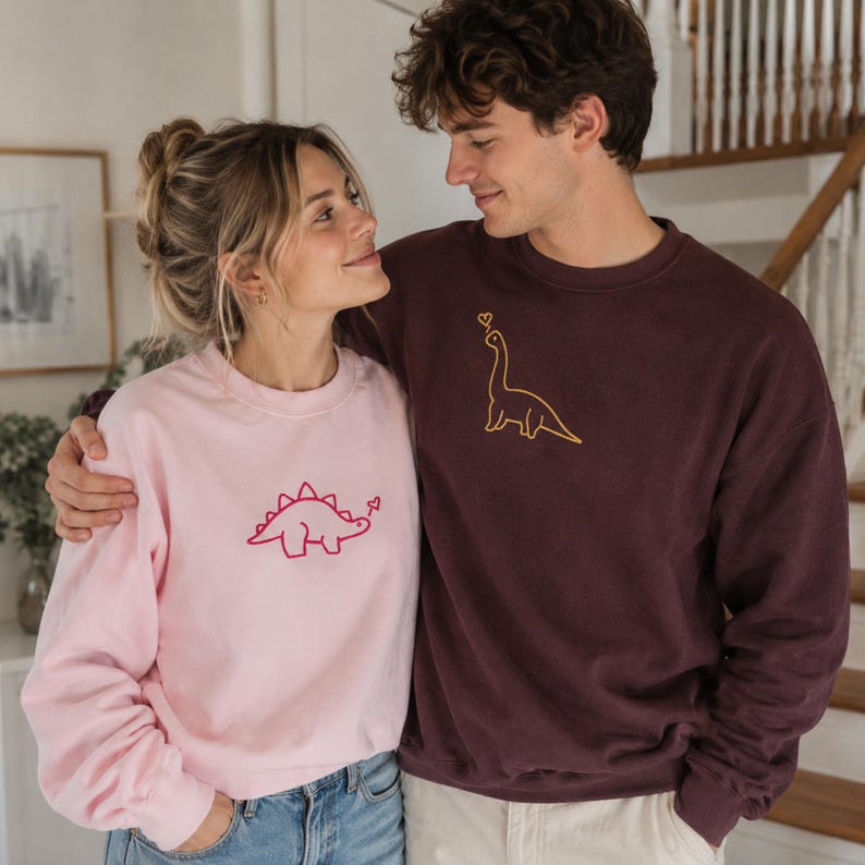 May include: A light pink crewneck sweatshirt featuring a red embroidered stegosaurus with a small heart, and a dark maroon crewneck sweatshirt featuring a yellow embroidered long-necked dinosaur with a small heart above its head.