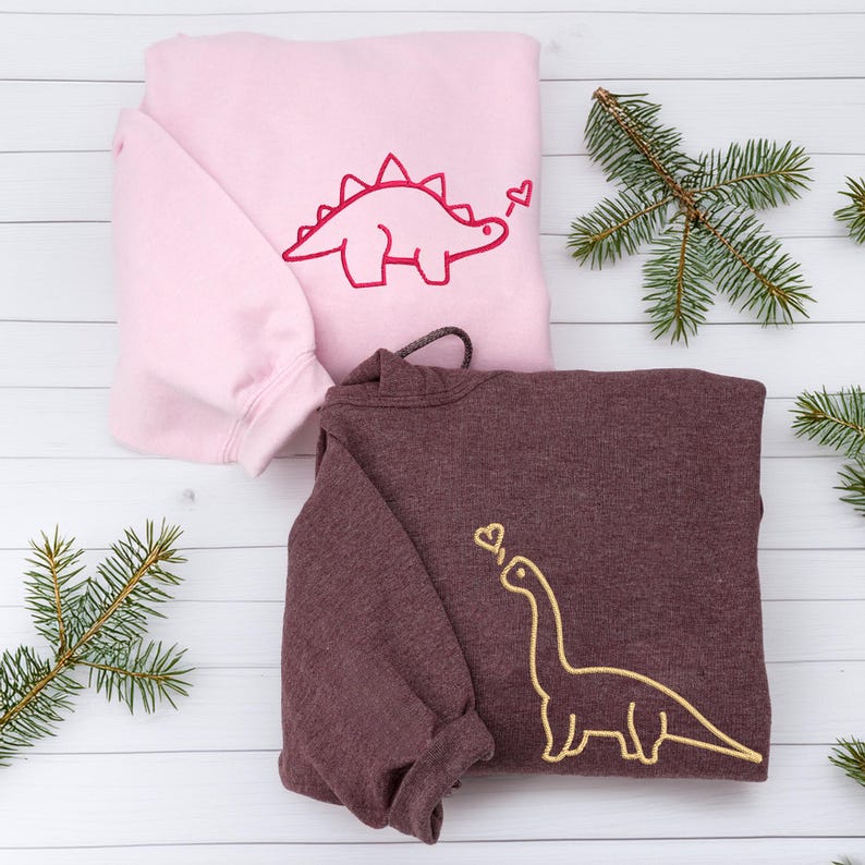 May include: A light pink sweatshirt featuring a dark pink embroidered stegosaurus with a heart, and a dark maroon sweatshirt featuring a gold embroidered long-necked dinosaur with a heart, both with long sleeves and ribbed cuffs.