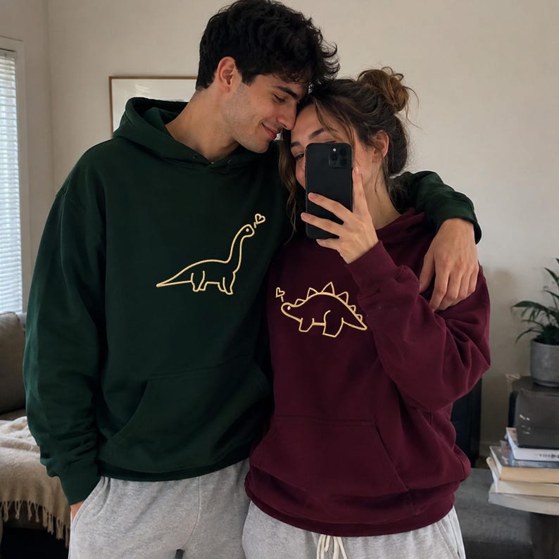 May include: A dark green hooded sweatshirt featuring a yellow line-art long-neck dinosaur and a maroon hooded sweatshirt featuring a yellow line-art spiked dinosaur, both with a small heart graphic above the dinosaur's head.