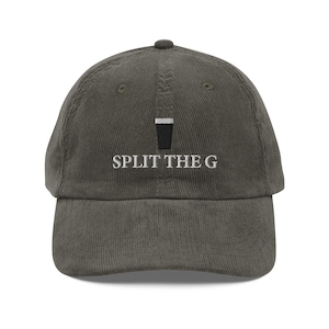 May include: A corduroy baseball cap in a muted olive green color. The cap features the embroidered text "SPLIT THE G" in white, with a black and white graphic of a pint glass above the text. The cap has a curved brim and a top button.