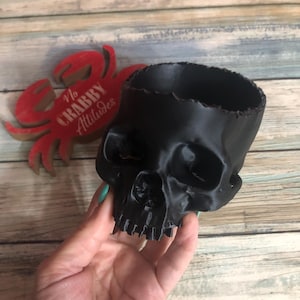 May include: A black skull-shaped planter with a jagged rim. The planter is held in a hand, and a red crab-shaped decoration with the text "No Crabby Attitudes" is in the background. The planter is made of a shiny material.