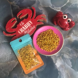 May include: A small, clear plastic bag filled with yellow bee pollen granules. The bag is on a pink and blue background. The text "No Crabby Attitudes" is visible on a red wooden crab.