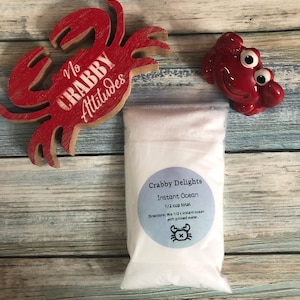 May include: A bag of "Crabby Delights Instant Ocean" with a blue label and crab graphic. A red crab-shaped wooden decoration with "No Crabby Attitudes" text and a red crab figurine are also present. The items are on a wood-grain surface.