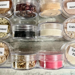 May include: A collection of small, clear plastic jars filled with various dried herbs, powders, and seeds. The jars are arranged in rows, with some labeled. Colors include brown, red, beige, and black. The image showcases a variety of natural ingredients.