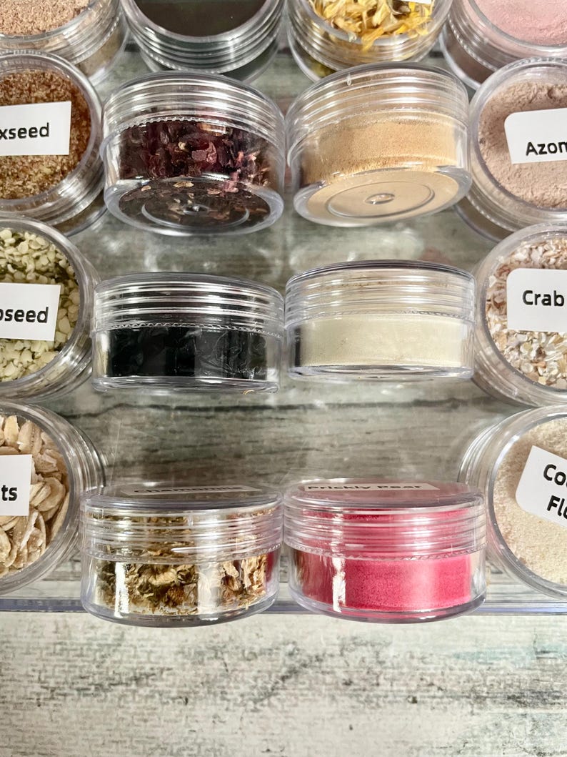 May include: A collection of small, clear plastic jars filled with various dried herbs, powders, and seeds. The jars are arranged in rows, with some labeled. Colors include brown, red, beige, and black. The image showcases a variety of natural ingredients.