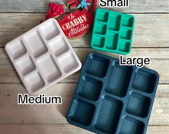 Könnte beinhalten: Drei quadratische Tabletts mit jeweils neun vertieften Fächern, beschriftet als Small in Grün, Medium in Hellrosa und Large in Dunkelblau. Eine rote Holzkrabben-Dekoration mit dem Text No CRABBY Attitudes befindet sich hinter den Tabletts.
