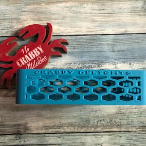 May include: A turquoise, honeycomb-patterned holder with the text "CRABBY DELIGHTS." A red crab-shaped decoration with the words "No CRABBY Attitudes" is also visible. The items are placed on a light blue and white wooden surface.