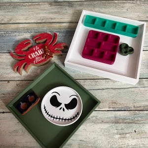 May include: A white square tray containing a teal five-compartment insert, a magenta nine-compartment insert, and a dark green textured piece. A green square tray holds a round white plate with a black skull face and a small rectangular dish.