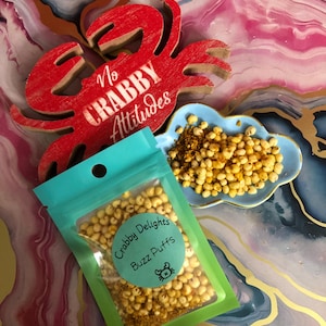 May include: A small, clear plastic bag filled with yellow and orange puffed cereal. The bag has a green and blue label that reads "Crabby Delights Buzz Puffs". A small red wooden crab with the text "No Crabby Attitudes" is in the background.