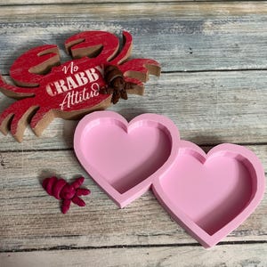 May include: A wooden red crab with the text "No Crabby Attitude" and a small brown crab figurine. Two pink heart-shaped molds and a small pink crab figurine are also present. The items are on a weathered wooden surface.
