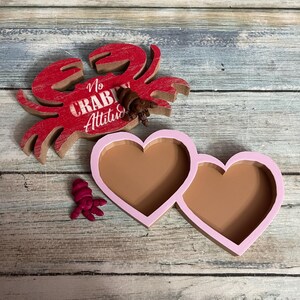 May include: A red wooden crab with the text "No Crab Attitude" and two pink heart-shaped trays with brown interiors. A small crab figurine and a small pink starfish figurine are also present. The items are on a weathered wood surface.