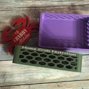 May include: A red crab-shaped decoration with the text "No Crabby Attitudes" next to a green honeycomb-patterned tray with the text "Home Crab Nutrition". A purple rectangular tray is also present.