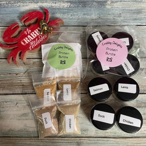 May include: An assortment of pet food products, including small bags of salmon, duck, lamb, and chicken protein, and small jars of the same. A red crab-shaped decoration with the text "No Crabby Attitudes" is also present.