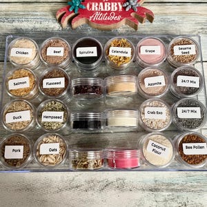 May include: A clear plastic tray holds small jars filled with various ingredients, including chicken, beef, spirulina, and sesame seeds. A red crab-shaped sign with the words "No Crabby Attitudes" sits above the jars.