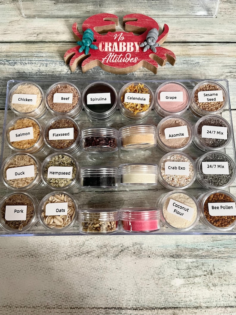 May include: A clear plastic tray holds small jars filled with various ingredients, including chicken, beef, spirulina, and sesame seeds. A red crab-shaped sign with the words "No Crabby Attitudes" sits above the jars.
