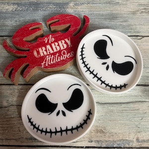 May include: Two white ceramic coasters with a black Jack Skellington face design and a red wooden crab with the text "No Crabby Attitudes". The coasters are round and have a slightly raised edge.