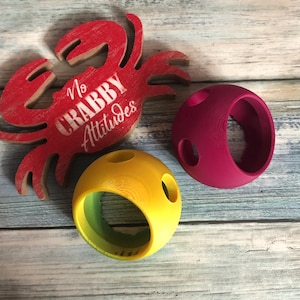 May include: A red wooden crab cutout with the white text No CRABBY Attitudes, accompanied by a yellow spherical object with circular cutouts and a magenta spherical object with circular cutouts, all featuring a smooth, matte finish.