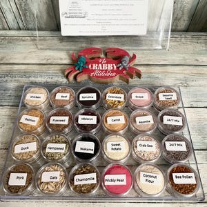 May include: A clear plastic tray filled with small, labeled containers of hermit crab nutrition. The labels include "Chicken," "Beef," "Spirulina," and more. A red crab-shaped decoration with the text "No Crabby Attitudes" sits above the tray.