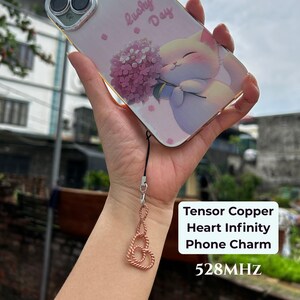 May include: A copper-colored metal phone charm featuring an infinity heart design attached to a smartphone case. The case displays a graphic of a white cat holding a purple flower bouquet with the text Lucky Day printed in pink script.
