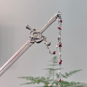 Könnte beinhalten: Ein silberner Haarstab mit einem schwertartigen Design. Der Stab hat ein dekoratives kreuzförmiges Element und eine Kette aus roten herzförmigen Perlen und kleinen weißen Perlen. Der Hintergrund ist ein weiches, verschwommenes Grün.
