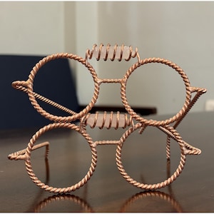 Puede incluir: Un soporte decorativo con forma de dos pares de gafas apiladas, construido con alambre de cobre trenzado. Cada puente presenta un cristal de cuarzo cilíndrico de color rosa pálido, envuelto en una pequeña espiral de alambre de cobre.