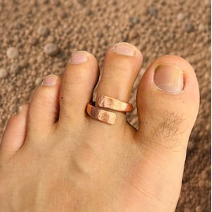May include: A copper-coloured toe ring with a hammered, textured finish. The ring features an open-ended, bypass design that allows for adjustment, with two slightly overlapping ends that wrap around the toe for a secure and comfortable fit.