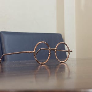 May include: Copper-colored wireframe glasses with round lenses and twisted wire detailing. The glasses are resting on a dark wooden surface, with a dark blue surface in the background.