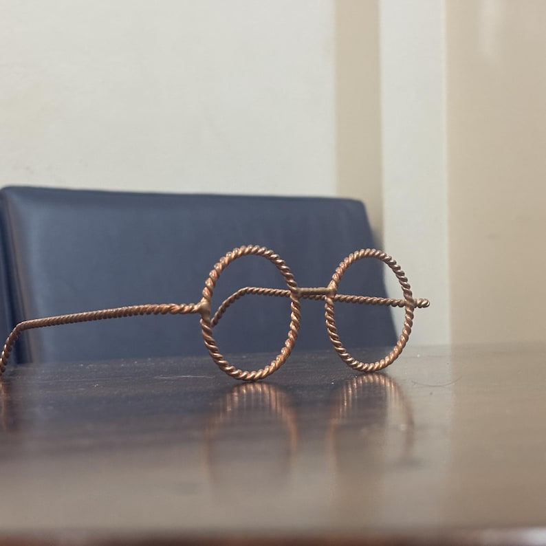 May include: Copper-colored wireframe glasses with round lenses and twisted wire detailing. The glasses are resting on a dark wooden surface, with a dark blue surface in the background.