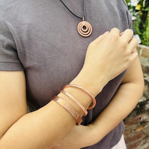 May include: A set of three copper-toned bracelets worn on the wrist, consisting of one smooth band, one twisted rope-style band, and one flat cuff. A matching circular pendant with a concentric rope-like design hangs on a black cord necklace.