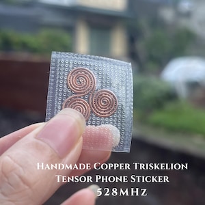 May include: A square clear adhesive sticker featuring a copper-colored triskelion design made of three connected spirals. The text on the sticker reads: HANDMADE COPPER TRISKELION TENSOR PHONE STICKER 528MHZ. The sticker has a textured dotted surface.